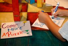 A child lettering a sign with a blue marker next to a sign that says "Come make a puzzle!: