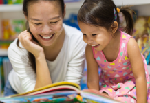 Asian mom and daughter smiling while looking at a children's book in a library setting.