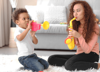 Female babysitter with long brown curly hair playing a yellow toy saxophone on the floor of a home next to a biracial male child playing a toy horn.